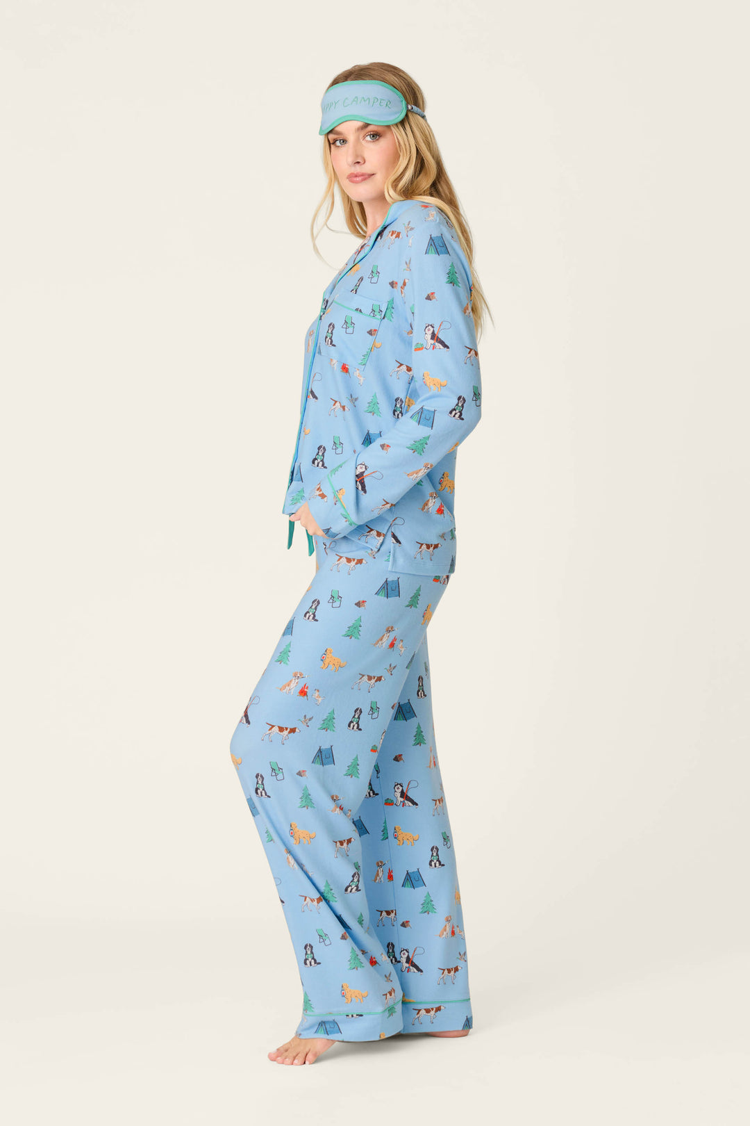 A woman stands barefoot in the P.J. Salvage Happy Campawr Dogs Camping Print 3-Piece PJ Set, featuring playful animal and tree designs in blue, with a matching blue sleep mask on her head. Her hands rest in her pajama pockets as she faces sideways against a light background.