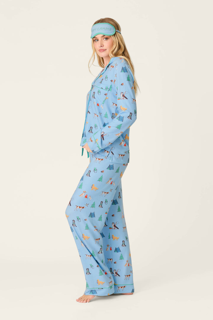 A woman stands barefoot in the P.J. Salvage Happy Campawr Dogs Camping Print 3-Piece PJ Set, featuring playful animal and tree designs in blue, with a matching blue sleep mask on her head. Her hands rest in her pajama pockets as she faces sideways against a light background.