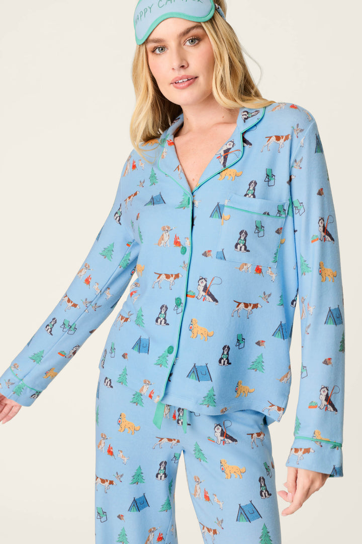 A woman smiles and poses against a neutral background, wearing the P.J. Salvage Happy Campawr Dogs Camping Print 3-Piece PJ Set, complete with a matching Happy Camper sleep mask.