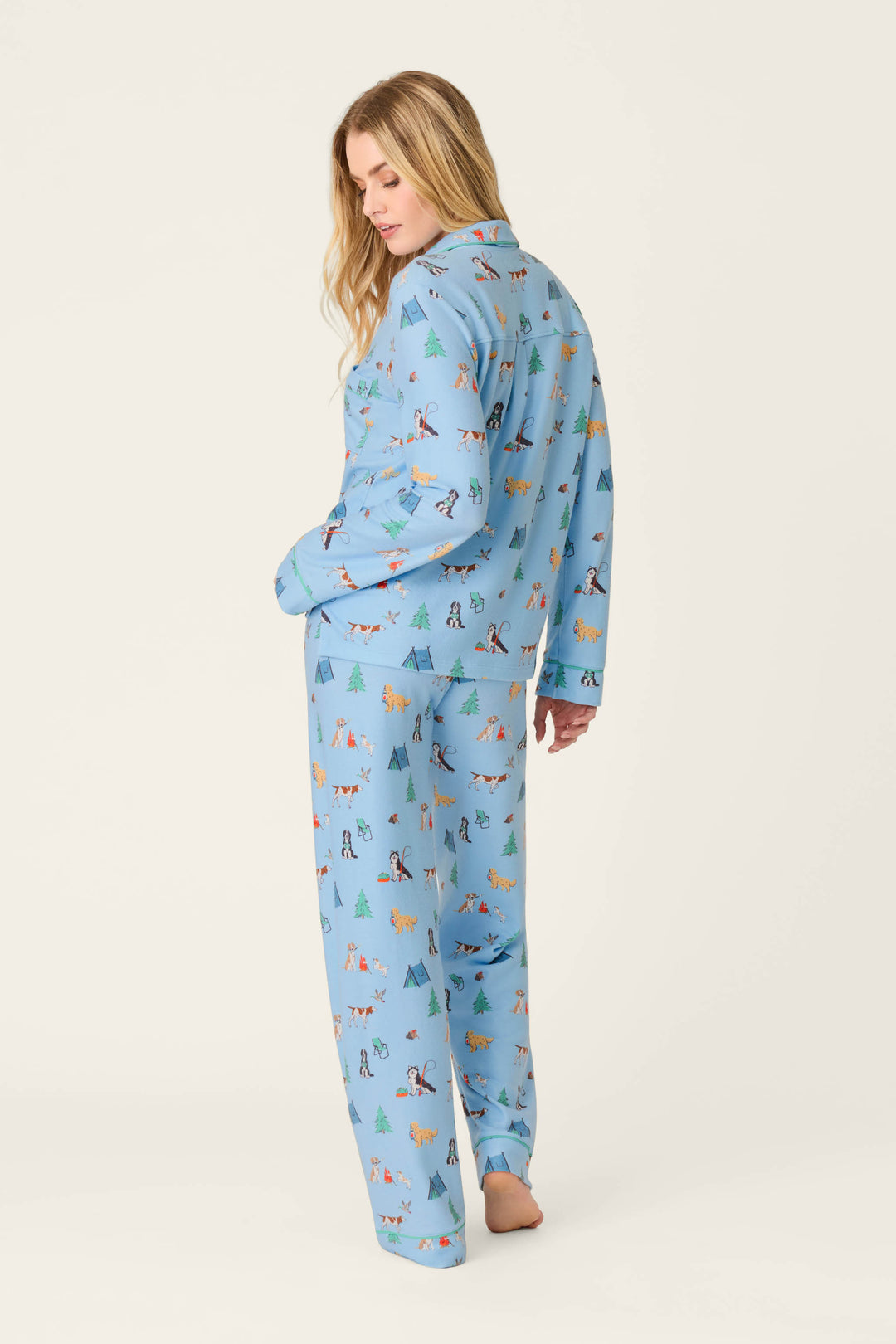 A person with long blonde hair models the P.J. Salvage Happy Campawr Dogs Camping Print 3-Piece PJ Set—light blue pajamas featuring colorful dog and tree prints—standing barefoot and glancing over their shoulder against a plain white background.