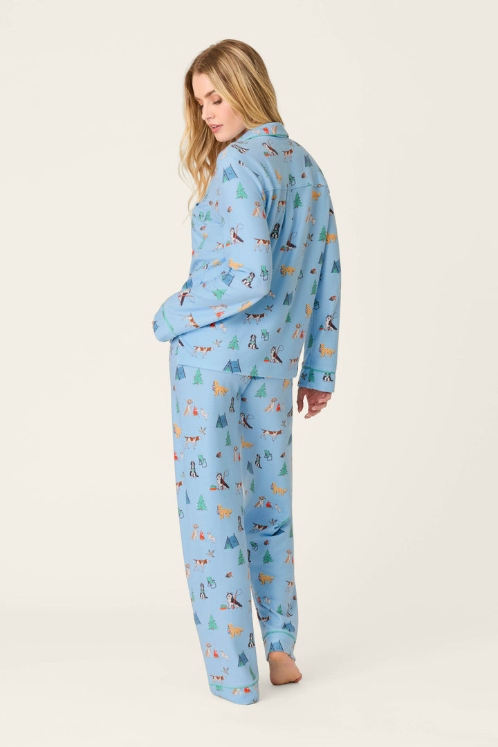 A person with long blonde hair models the P.J. Salvage Happy Campawr Dogs Camping Print 3-Piece PJ Set—light blue pajamas featuring colorful dog and tree prints—standing barefoot and glancing over their shoulder against a plain white background.