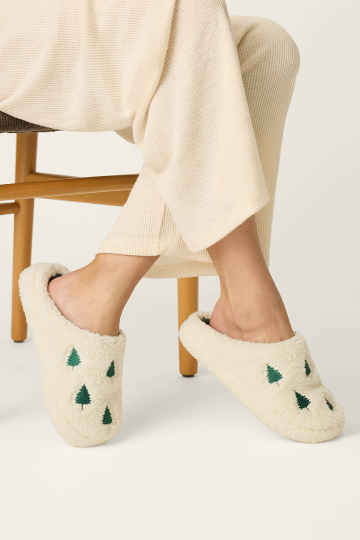 A person sits on a wooden chair wearing cream-colored loungewear and cozy white P.J. Salvage Tree Print Faux Sherpa Slippers adorned with small green tree patterns.