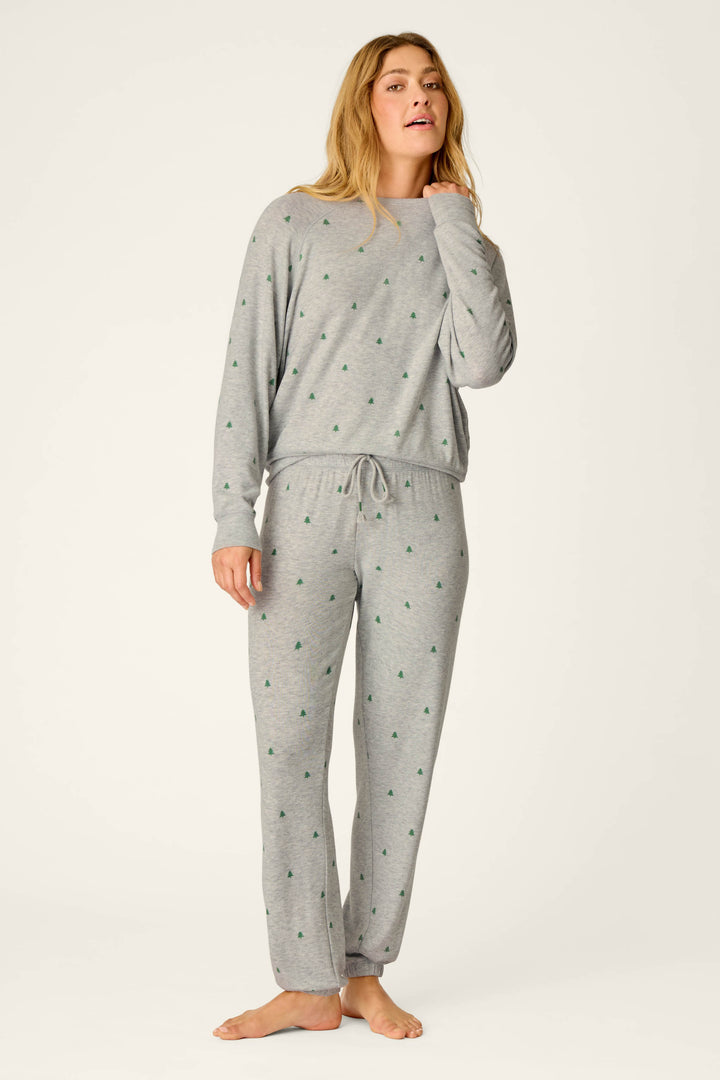 A woman stands barefoot against a plain light background, wearing the P.J. Salvage Happy Campawr Tree Print Long Sleeve & Pants Lounge Set—a matching grey pajama set featuring small green tree patterns, long sleeves, and drawstring pants.