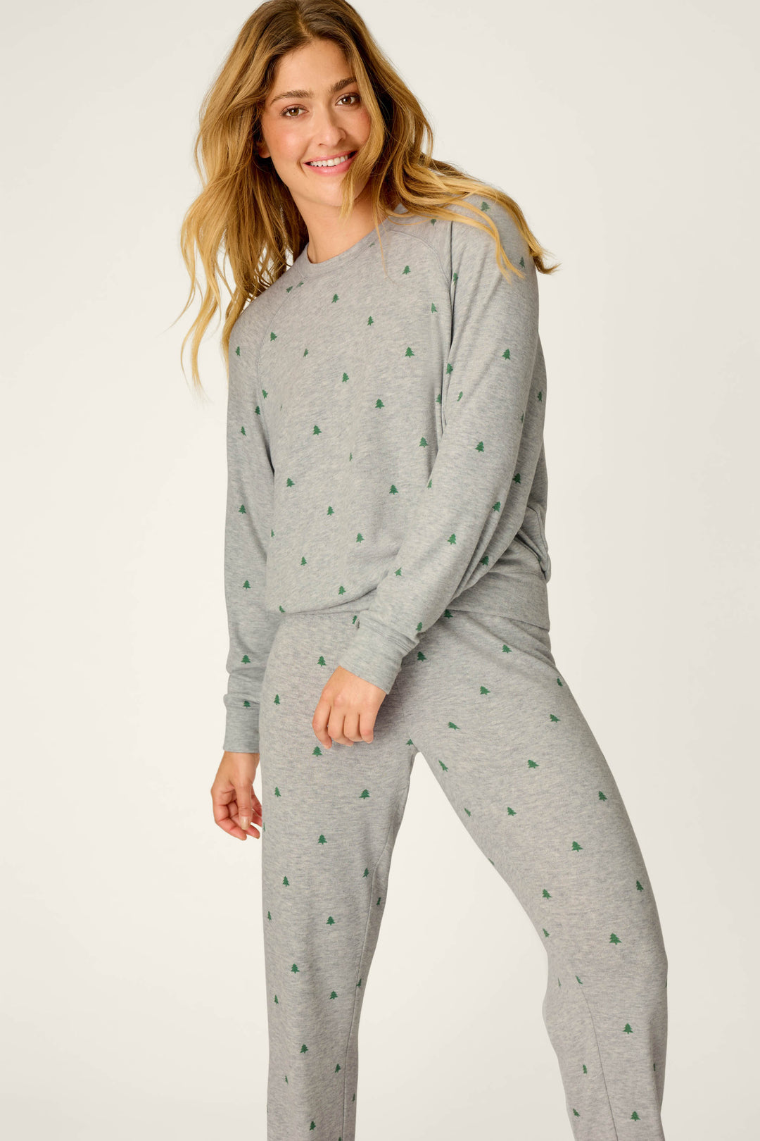A person with long hair smiles while wearing the P.J. Salvage Happy Campawr Tree Print Long Sleeve & Pants Lounge Set, featuring a matching grey design adorned with small green tree prints, standing against a plain light background.