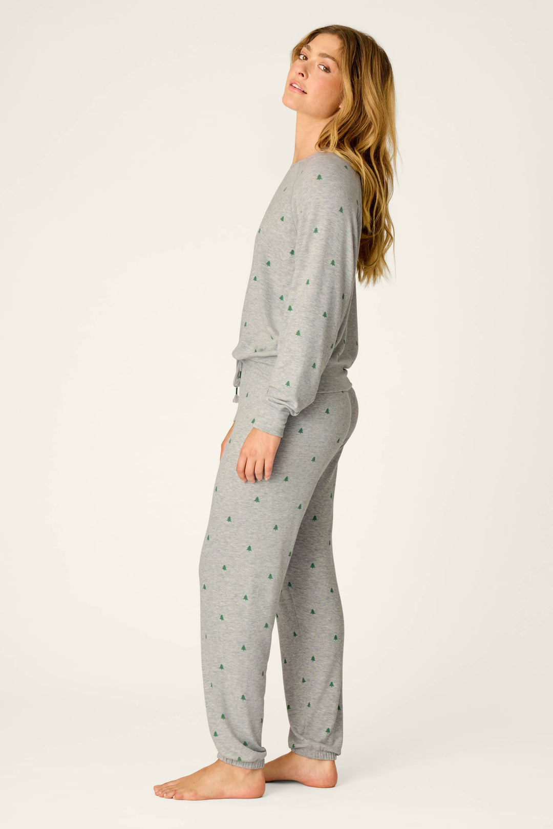 A woman stands barefoot and sideways against a plain light background, wearing the P.J. Salvage Happy Campawr Tree Print Long Sleeve & Pants Lounge Set in grey, featuring small green tree patterns, as she looks slightly over her shoulder.