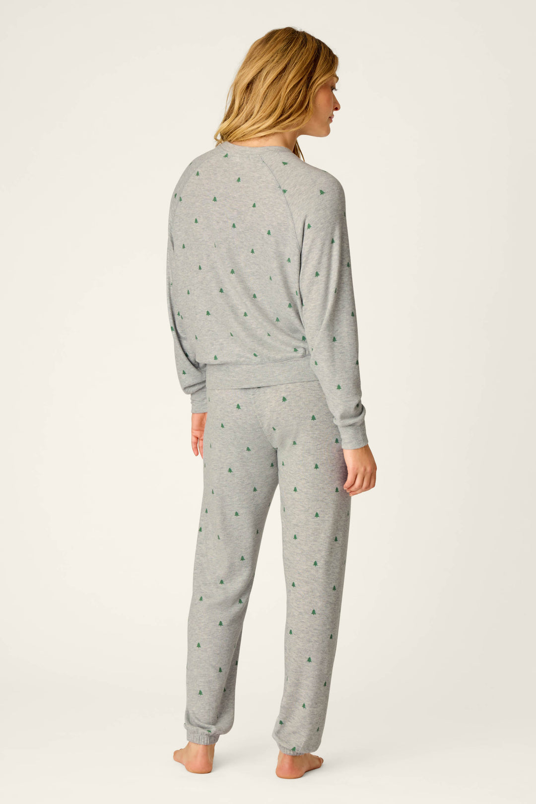 A person with long blonde hair, seen from behind, models the P.J. Salvage Happy Campawr Tree Print Long Sleeve & Pants Lounge Set—a matching light gray pajama set with small green tree prints—standing against a plain off-white background.