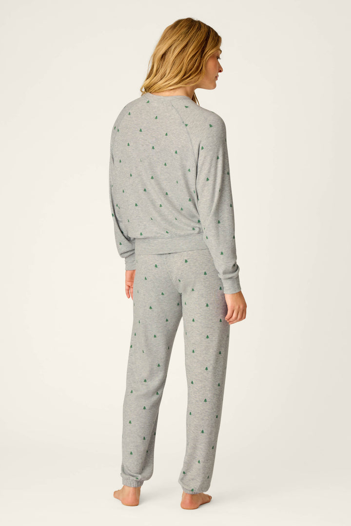 A person with long blonde hair, seen from behind, models the P.J. Salvage Happy Campawr Tree Print Long Sleeve & Pants Lounge Set—a matching light gray pajama set with small green tree prints—standing against a plain off-white background.