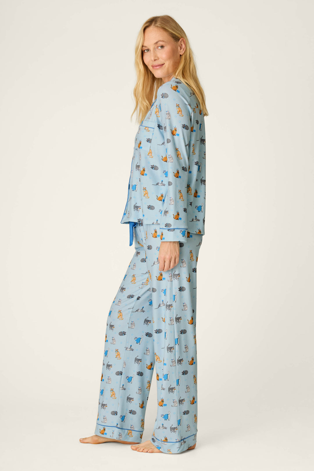 A woman with long blonde hair stands barefoot and smiling, facing slightly to the side against a plain light background, wearing the P.J. Salvage Hanukkatz Paws for Celebration Cat Print PJ Set.
