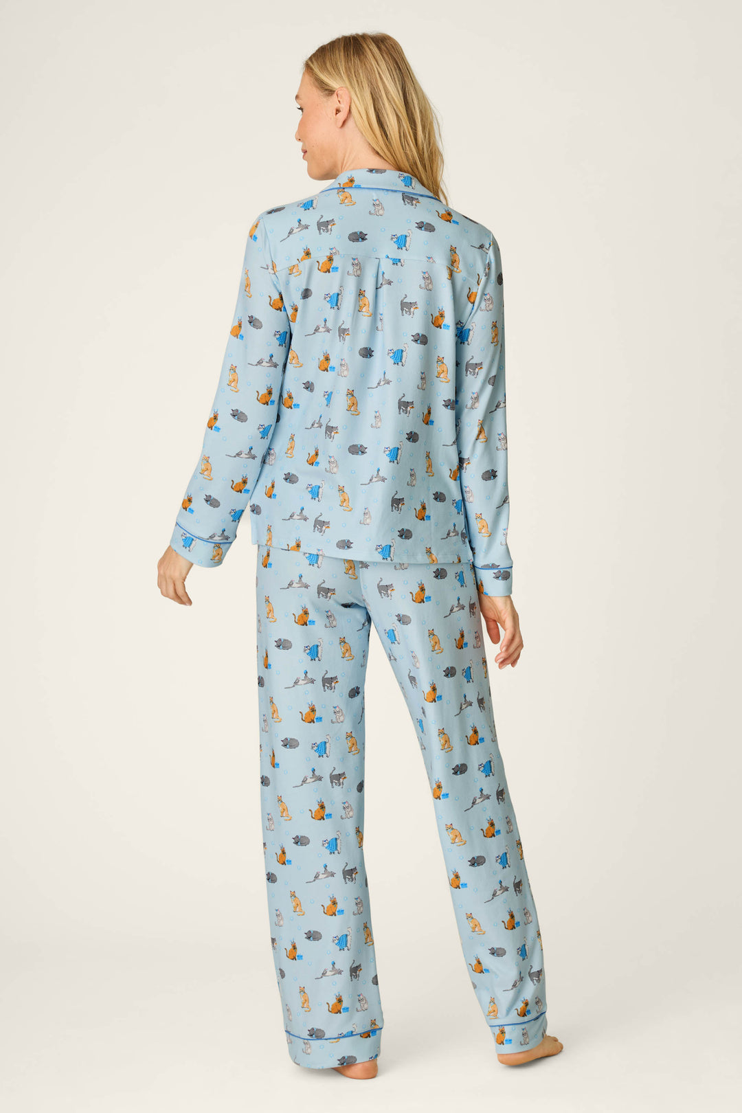 A woman stands barefoot, facing away, wearing the Hanukkatz Paws for Celebration Cat Print PJ Set by P.J. Salvage. The light blue pajama top features a playful cat print, a collar, and long sleeves, while the matching pants are loose-fitting. The background is a plain off-white.