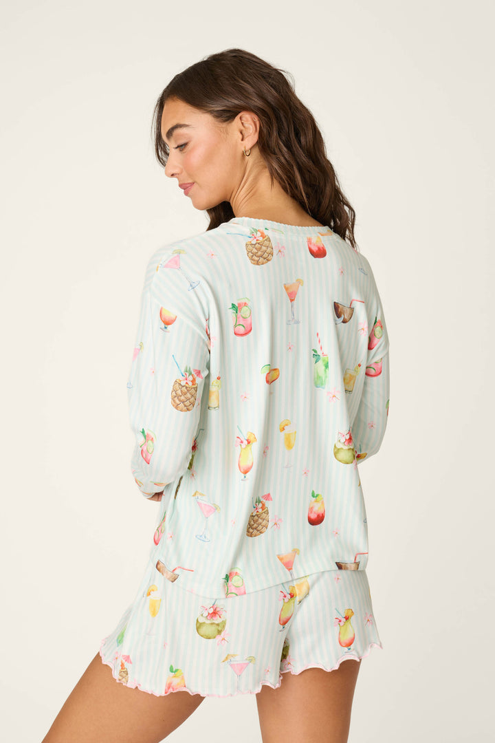 A woman with long brown hair wears the P.J. Salvage "It's Five O'clock Somewhere Cocktail Print" long sleeve top, featuring colorful cocktail illustrations, as she turns to show the back against a plain background.