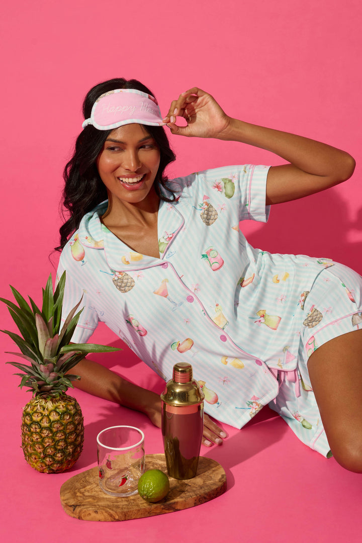 A woman in the P.J. Salvage It's Five O'clock Somewhere Cocktail Print 3-Piece PJ Set and a pink sleep mask poses on a pink background with cocktail-themed props.