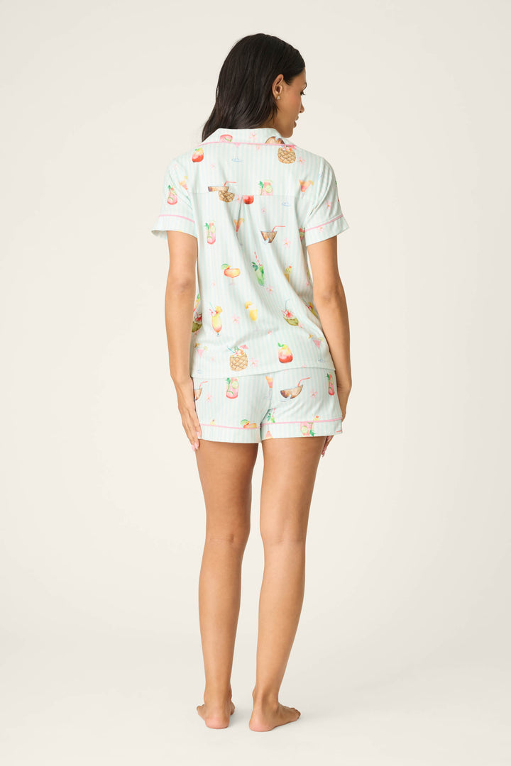A woman stands barefoot, facing away, in the P.J. Salvage It's Five O'clock Somewhere Cocktail Print 3-Piece PJ Set featuring light blue fabric with colorful fruit and drink motifs against a plain off-white background.