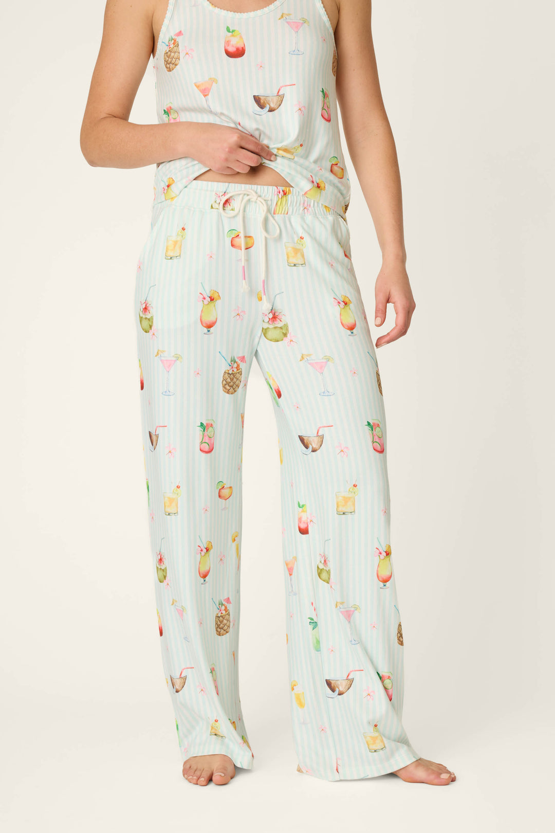 A person stands barefoot against a plain background wearing P.J. Salvage's It's Five O'clock Somewhere Cocktail Print Open Leg Pants, featuring a colorful cocktail print, drawstring waist, and relaxed fit.