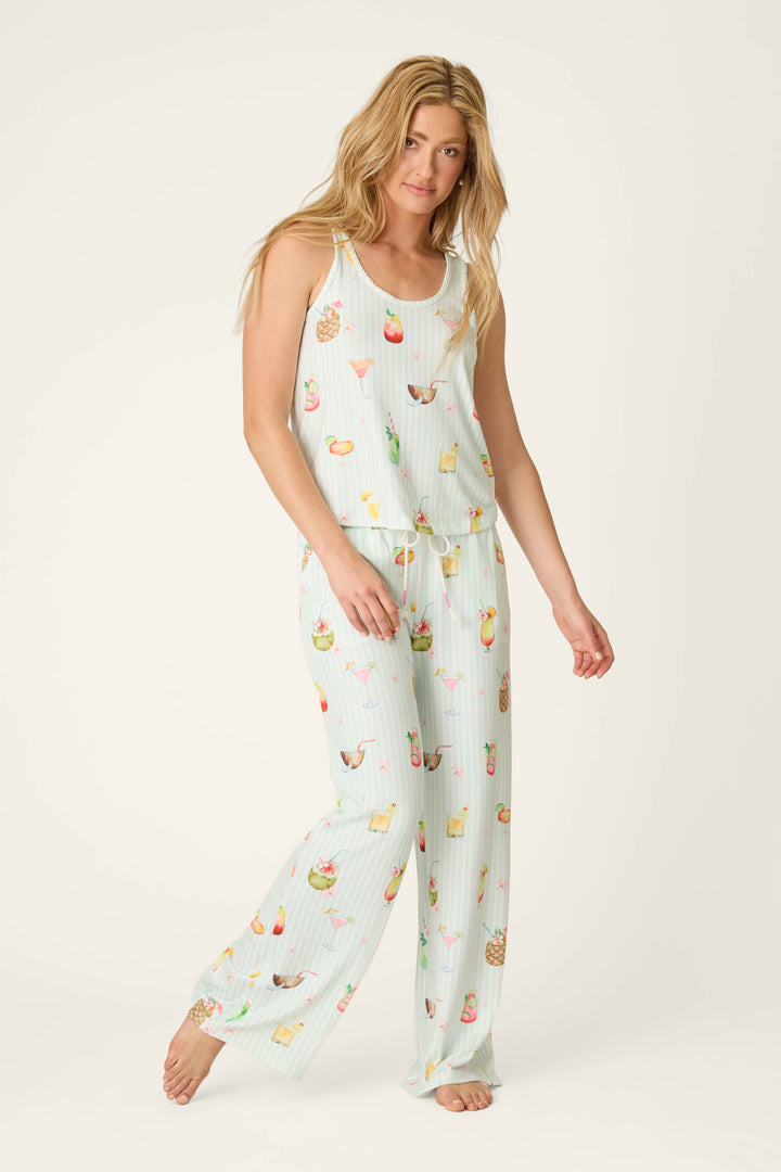 A woman with long blonde hair models the P.J. Salvage It's Five O'clock Somewhere Cocktail Print Open Leg Pants, featuring colorful tropical drink motifs, along with a matching tank top, standing barefoot against a light background.