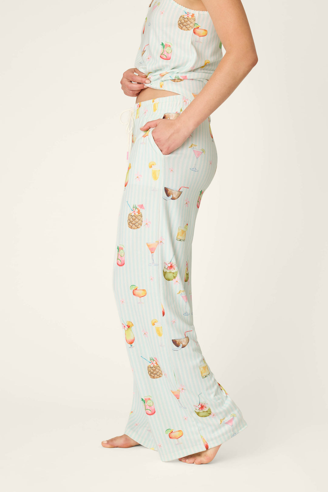 A person models P.J. Salvage's It's Five O'clock Somewhere Cocktail Print Open Leg Pants—light blue pajama bottoms featuring a vibrant tropical drink and fruit motif, shown barefoot with only the lower body and part of the torso visible.