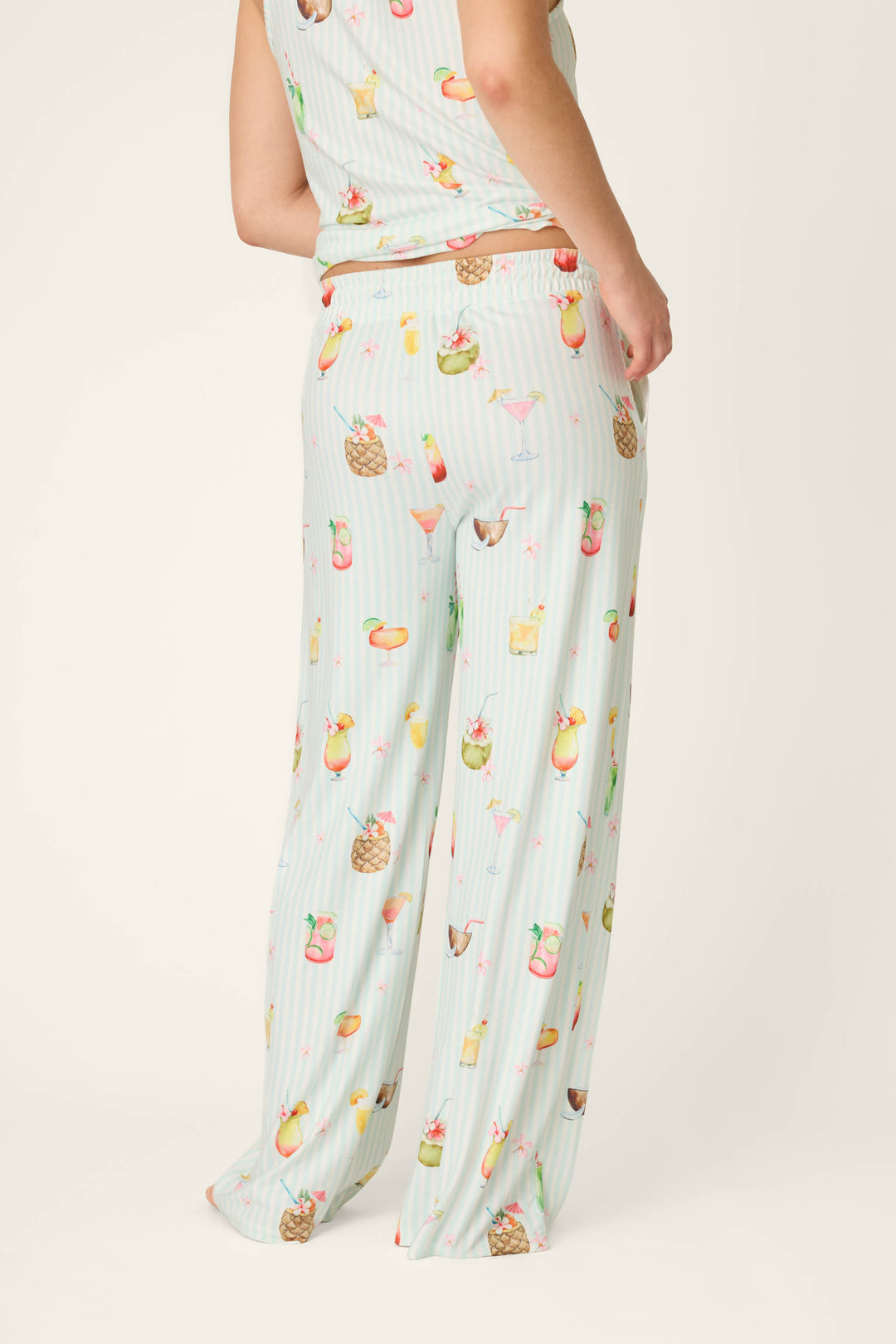 A person stands with their back partly turned, wearing P.J. Salvage It's Five O'clock Somewhere Cocktail Print Open Leg Pants, featuring a playful colorful cocktail and fruit pattern against a light, plain background.
