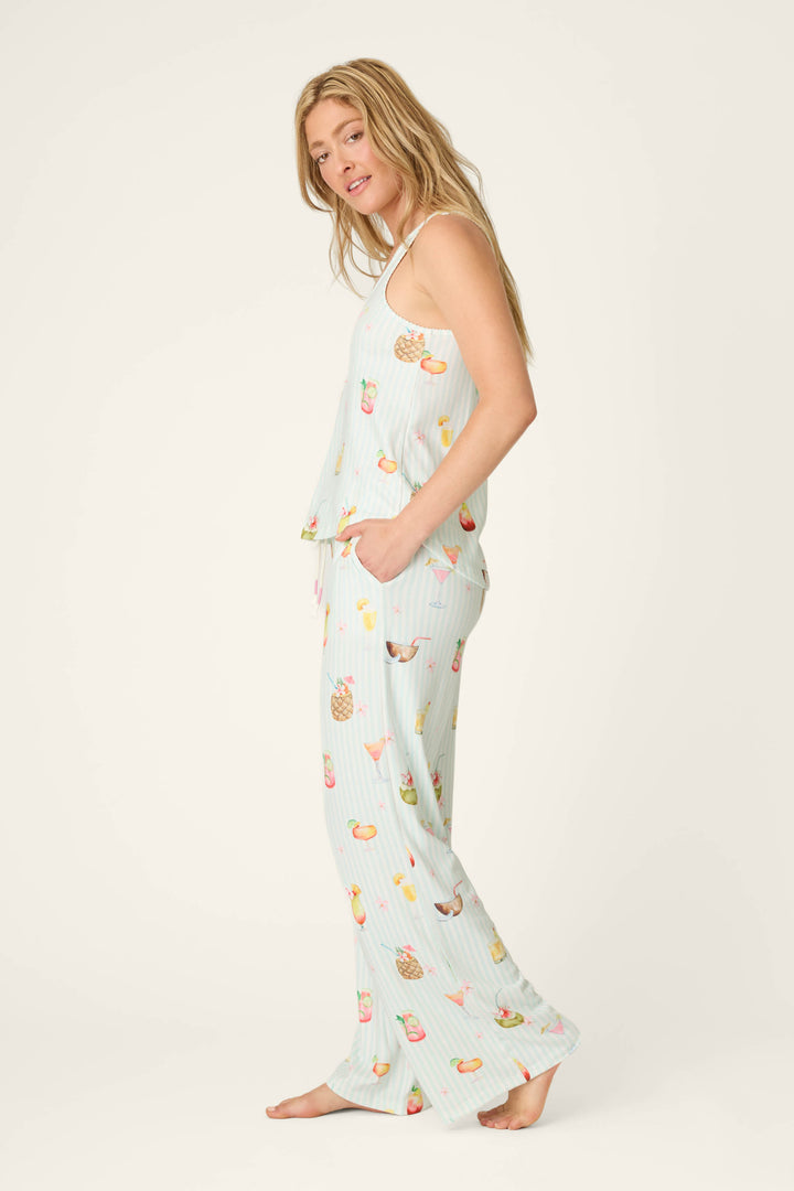 A woman stands barefoot in the P.J. Salvage It's Five O'clock Somewhere Cocktail Print Long PJ Set, featuring a sleeveless top and wide-leg pants with pockets in light blue fabric and colorful cocktail prints, facing sideways toward the camera.