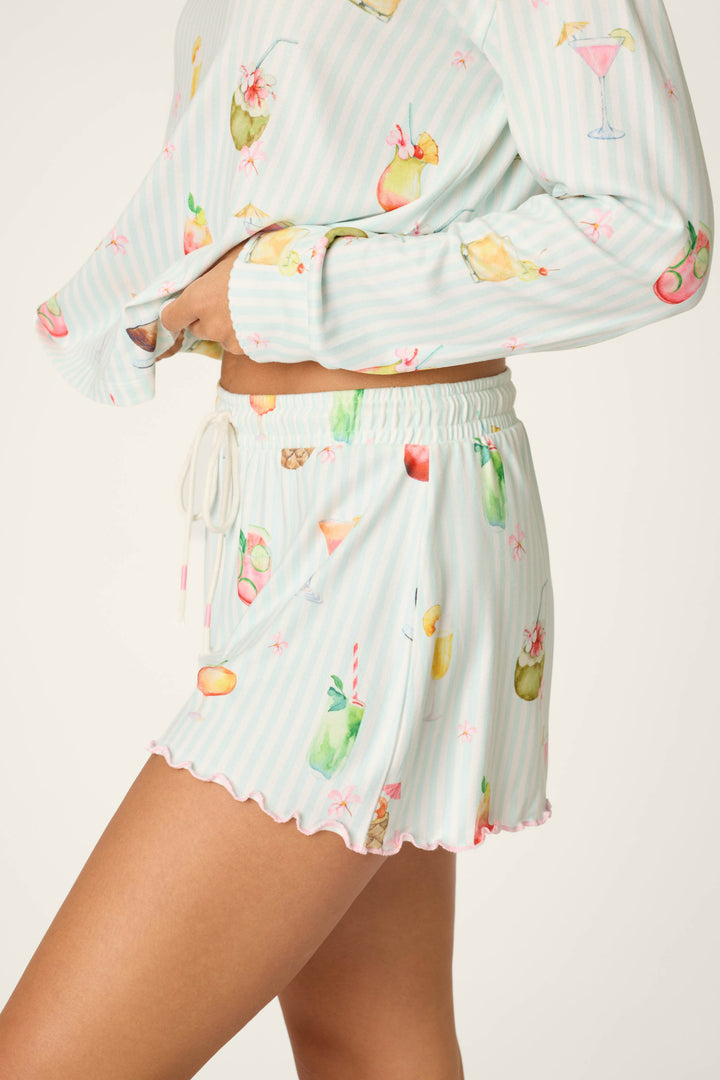 A person models P.J. Salvage's "It's Five O'clock Somewhere Cocktail Print Shorts"—pastel blue and white striped, colorful cocktail illustrations, relaxed fit, drawstring waist shorts and matching long-sleeve top. Only torso and legs are shown.
