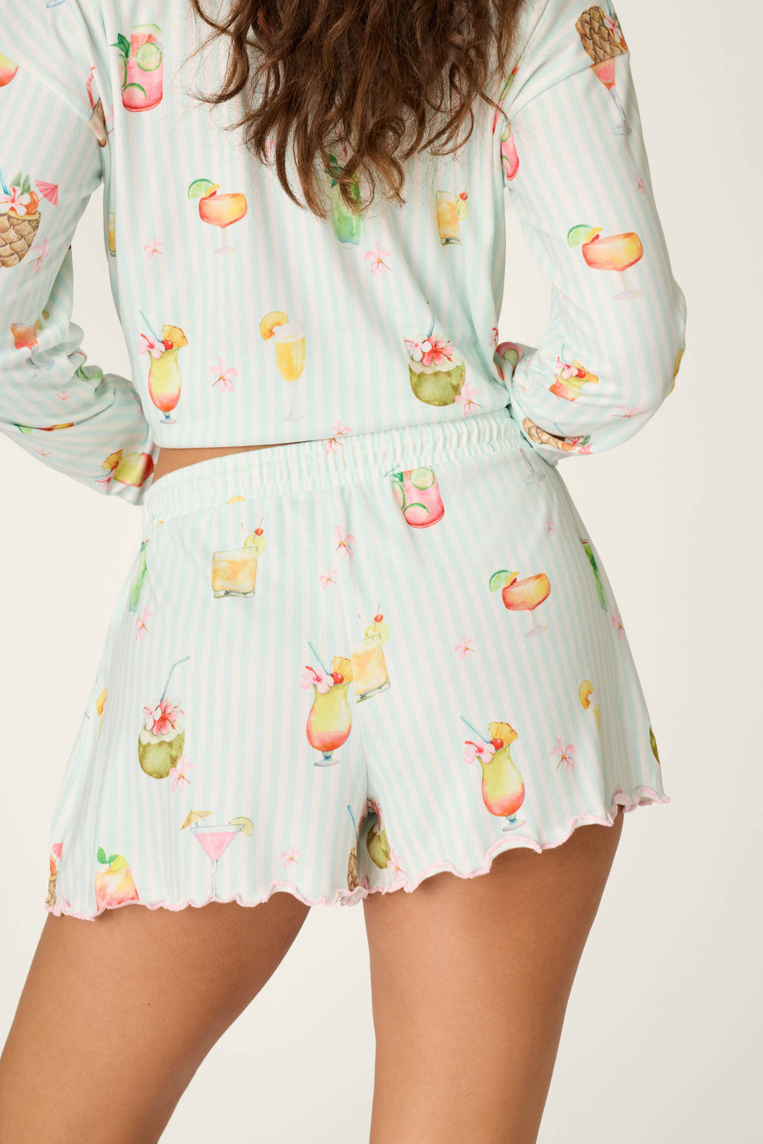 A woman is shown from the waist down, wearing P.J. Salvage's It's Five O'clock Somewhere Cocktail Print Shorts with a matching top featuring colorful tropical drinks and fruit prints, her long wavy hair over her shoulder and one hand on her hip.