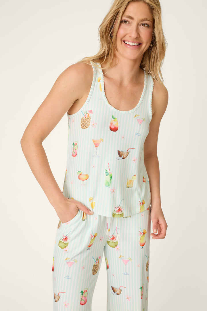 A woman smiles with one hand in her pocket, wearing the P.J. Salvage It's Five O'clock Somewhere Cocktail Print Tank Top—a sleeveless, light-colored pajama top featuring colorful tropical drinks and fruits.