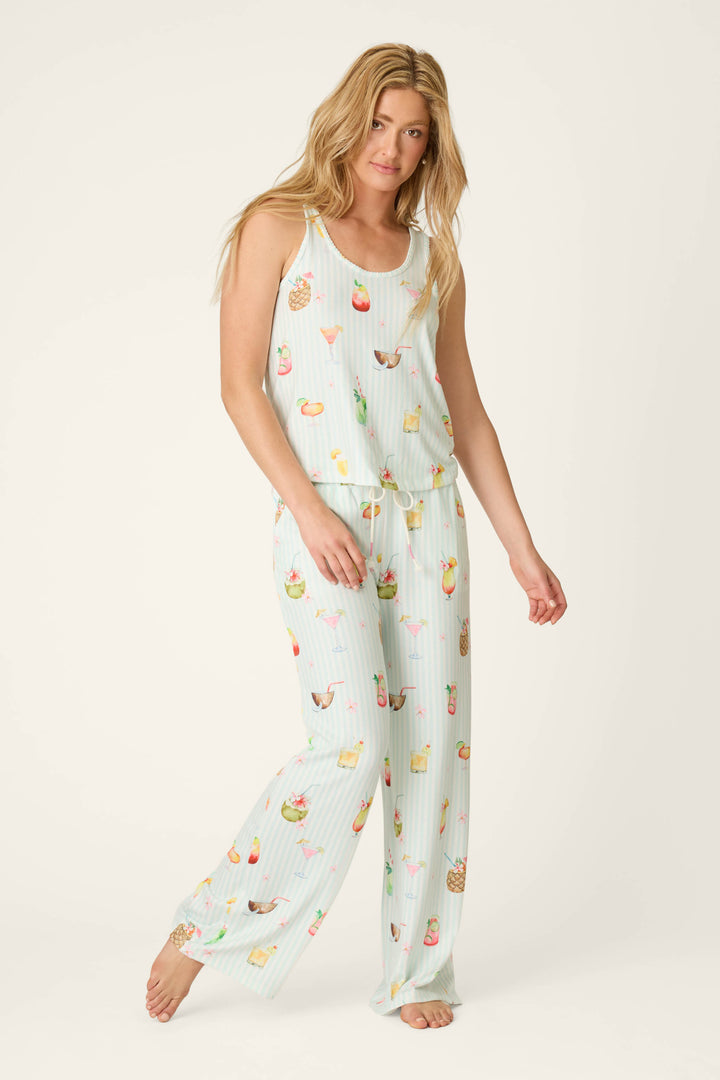 A woman with long blonde hair models the P.J. Salvage It's Five O'clock Somewhere Cocktail Print Tank Top, a light blue sleeveless pajama top with colorful tropical drink prints, standing barefoot against a neutral background.