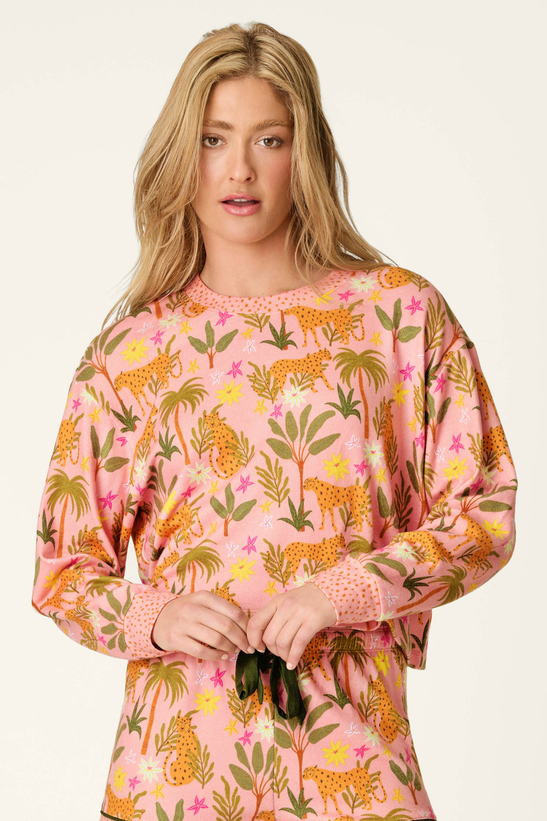 A woman with long blonde hair models the P.J. Salvage In the Jungle Leopard Safari Long Sleeve Top, a cropped, vibrant jungle print with tigers, plants, and flowers in green, yellow, and orange tones against a plain light background.