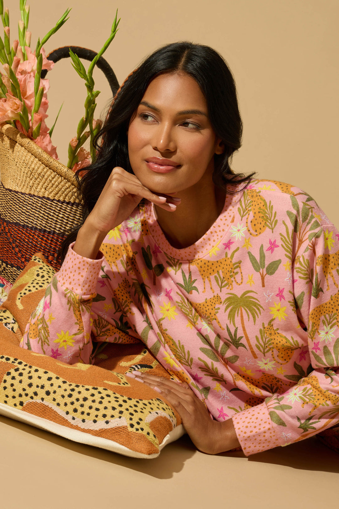 A woman with long black hair wears the P.J. Salvage "In the Jungle Leopard Safari" long-sleeve top, featuring vibrant animal and plant prints, as she leans on cheetah print pillows beside a woven flower basket against a tan background.