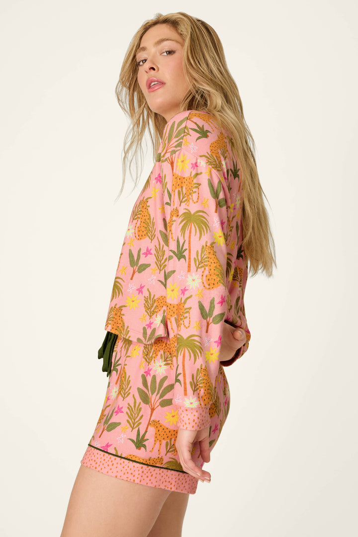 A woman with long blonde hair models the P.J. Salvage "In the Jungle Leopard Safari" long sleeve top, featuring a vibrant jungle print with cheetahs, leopards, leaves, and flowers. She stands sideways, looking back at the camera.