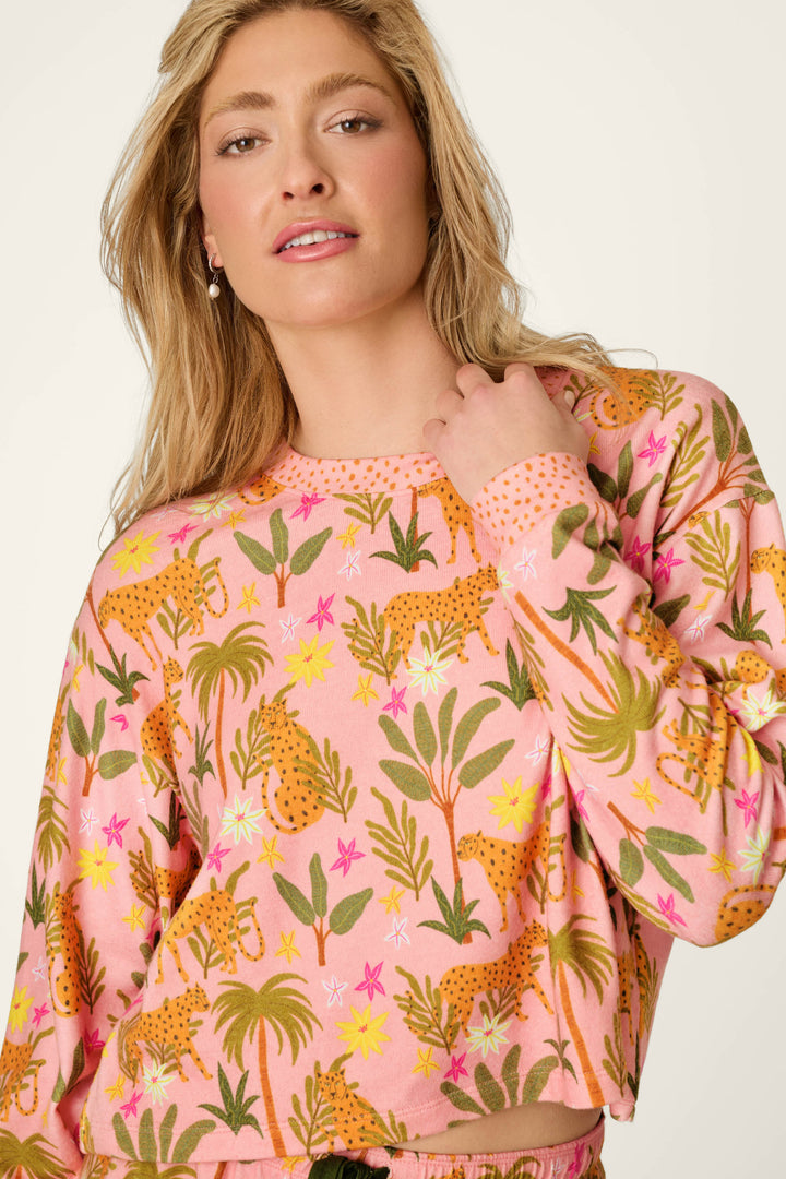 A person with long blonde hair wears the P.J. Salvage In the Jungle Leopard Safari Long Sleeve Top, facing the camera and holding their shirt collar. The pink cropped top features a jungle print with leaves, flowers, and leopards.