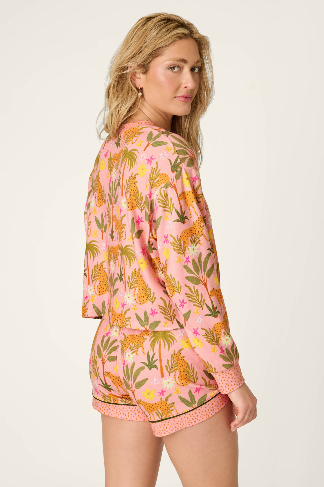 A woman with long blonde hair models the P.J. Salvage In the Jungle Leopard Safari Long Sleeve Top, featuring a colorful cheetah and floral print in pink, as she looks over her shoulder against a plain light background.