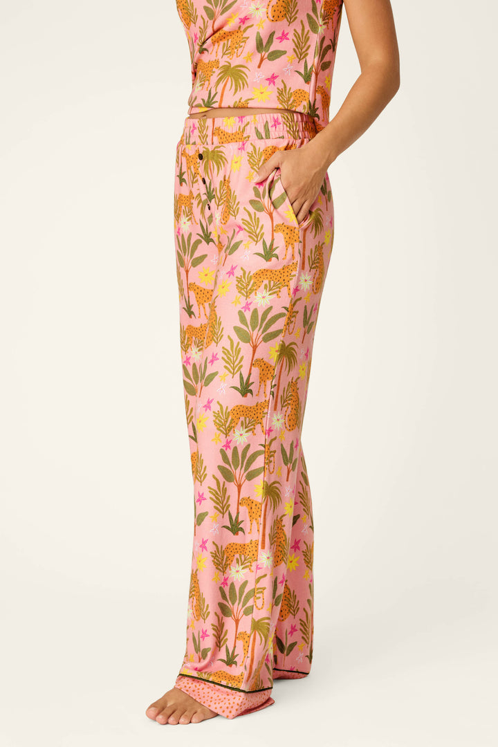 A person stands barefoot in P.J. Salvage’s In the Jungle Leopard Safari Open Leg Pants, featuring a pink base with a colorful jungle print and cheetahs, hand casually in one pocket. Upper body partially visible.