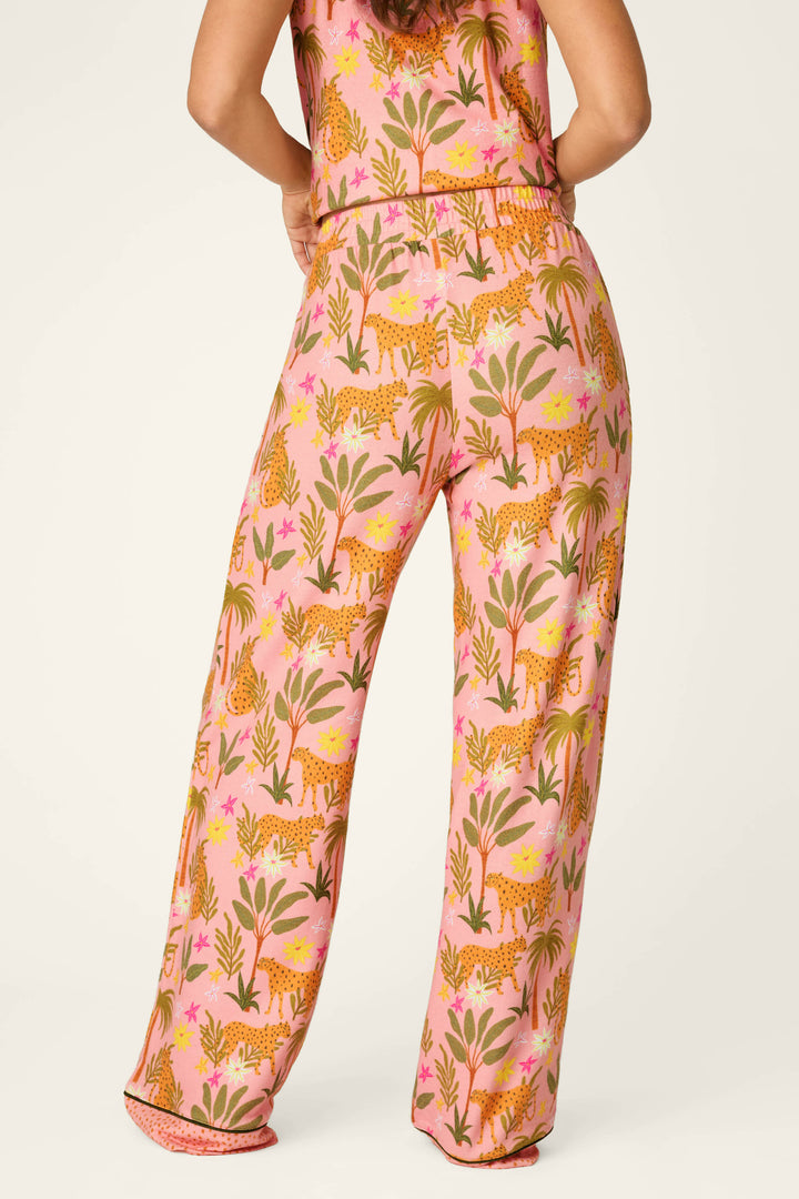 A person faces away, wearing P.J. Salvage's In the Jungle Leopard Safari Open Leg Pants featuring a colorful print of leopards and green plants. The background is plain and light-colored.