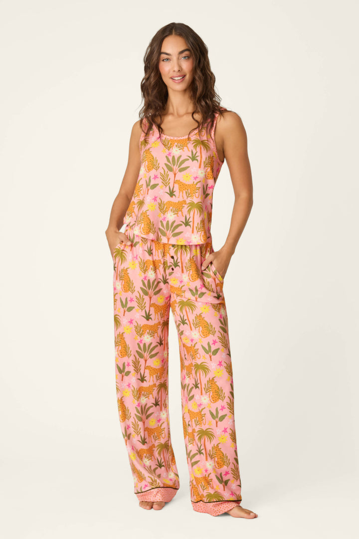 A woman smiles barefoot, wearing the P.J. Salvage "In the Jungle Leopard Safari Long PJ Set," a soft jersey sleepwear set in blush pink with a colorful leopard and plant print. The set features a sleeveless top, wide-leg pants, and handy pockets.