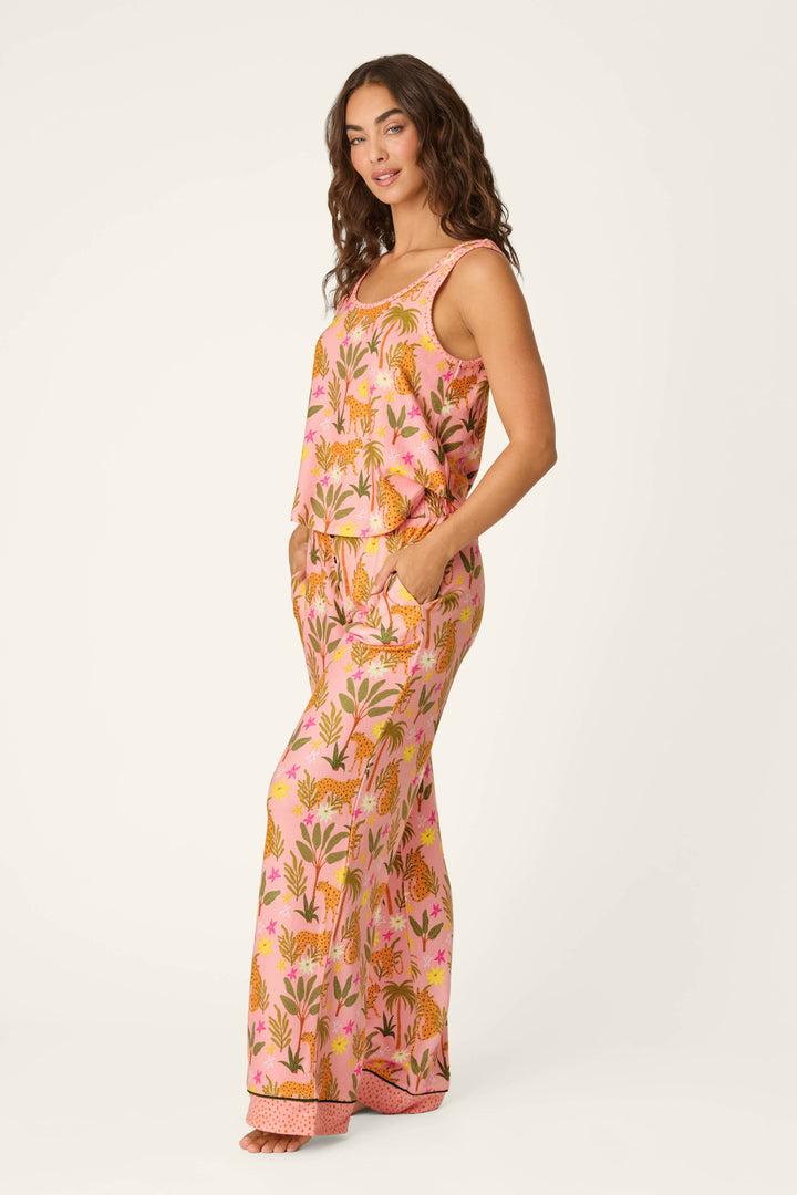 A woman with long curly hair stands barefoot against a plain light background, smiling slightly in the P.J. Salvage "In the Jungle Leopard Safari Long PJ Set," featuring a blush pink color with a vibrant botanical and leopard print.
