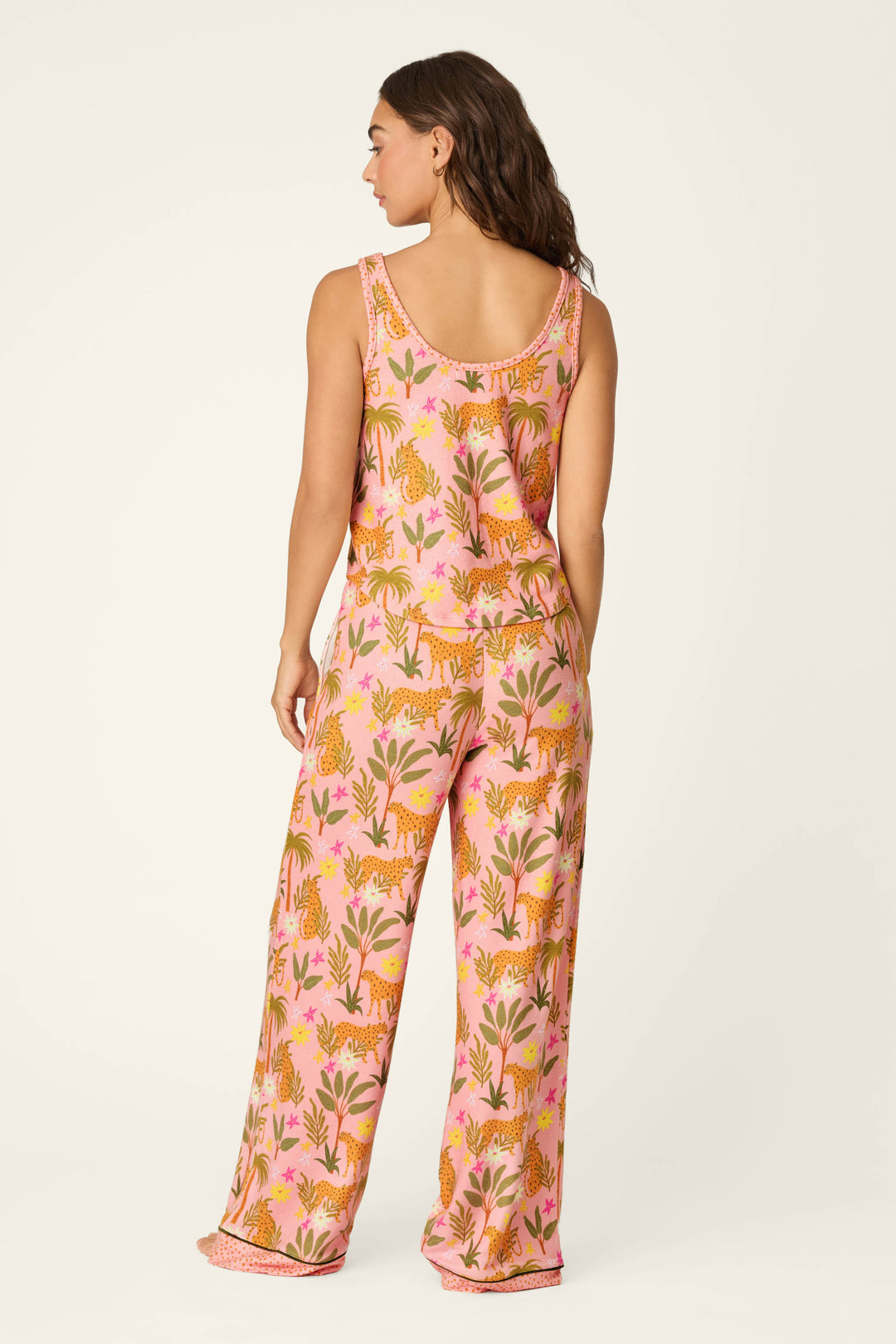 A woman faces away, modeling the P.J. Salvage In the Jungle Leopard Safari Long PJ Set—a sleeveless, loose-fit jersey pajama set in a vibrant pink, orange, and green jungle pattern—against a plain off-white background.