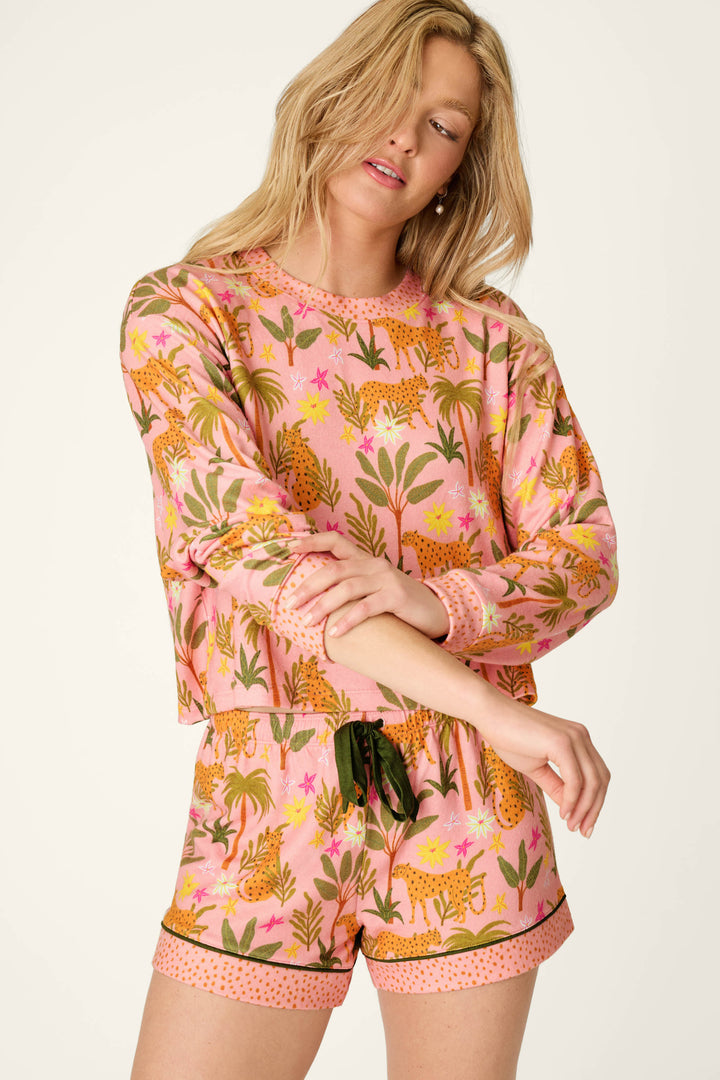 A woman with blonde hair models the P.J. Salvage In the Jungle Leopard Safari Short PJ Set, featuring an oversized peachy jersey silhouette with a bold leopard and floral print, long-sleeve top, and shorts as she poses with arms loosely crossed.