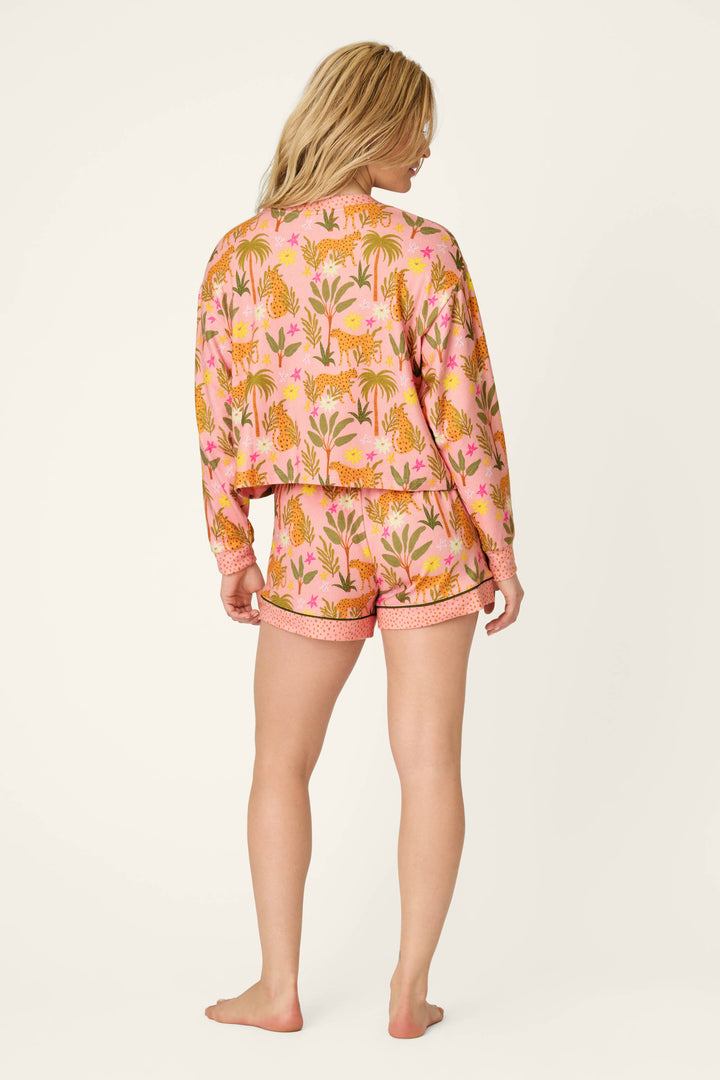 A woman stands barefoot facing away in the P.J. Salvage In the Jungle Leopard Safari Short PJ Set—a peachy, oversized jersey pajama set with a vibrant leopard and plant pattern, relaxed long-sleeve top, and shorts. Her blonde hair is loose against an off-white background.