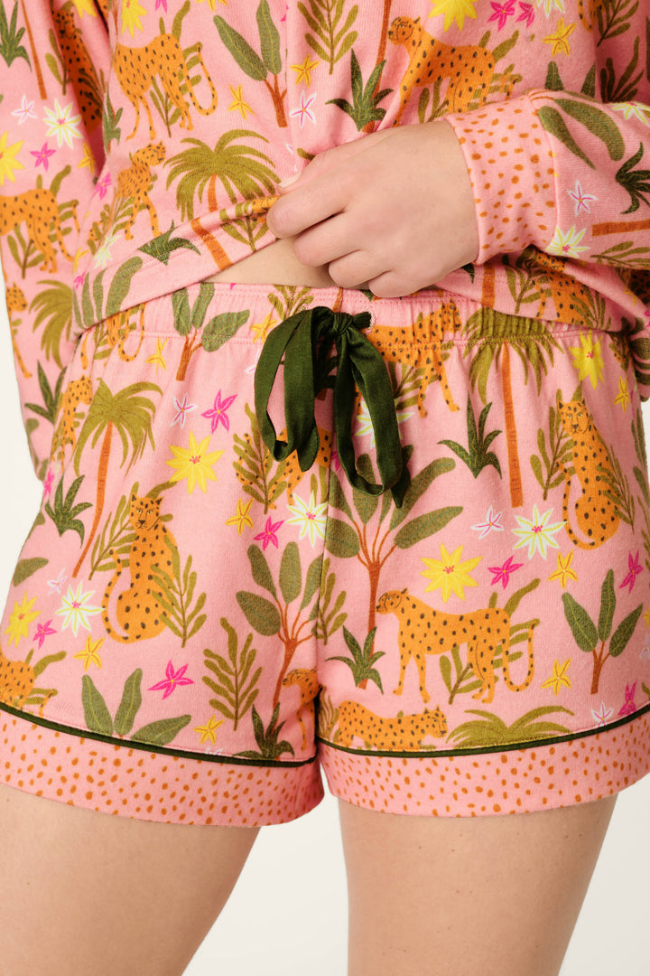 A person wearing P.J. Salvage In the Jungle Leopard Safari Shorts, featuring a peachy jersey fabric with cheetahs, green leaves, pink and yellow flowers, black trim at the hem, and a green ribbon drawstring.