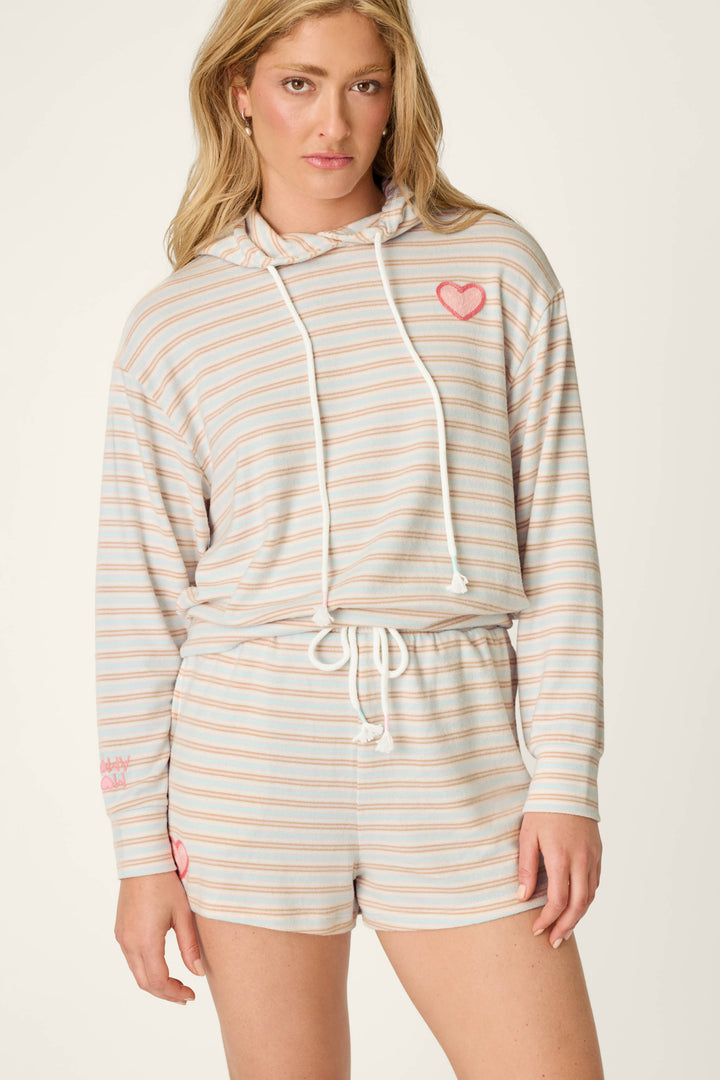 A woman wears the P.J. Salvage J'Adore Amour Sweetheart Stripes Hoodie and matching drawstring shorts, featuring cozy pastel stripes and charming red heart accents on the chest and shorts—an ideal loungewear set for relaxing in style against a light background.