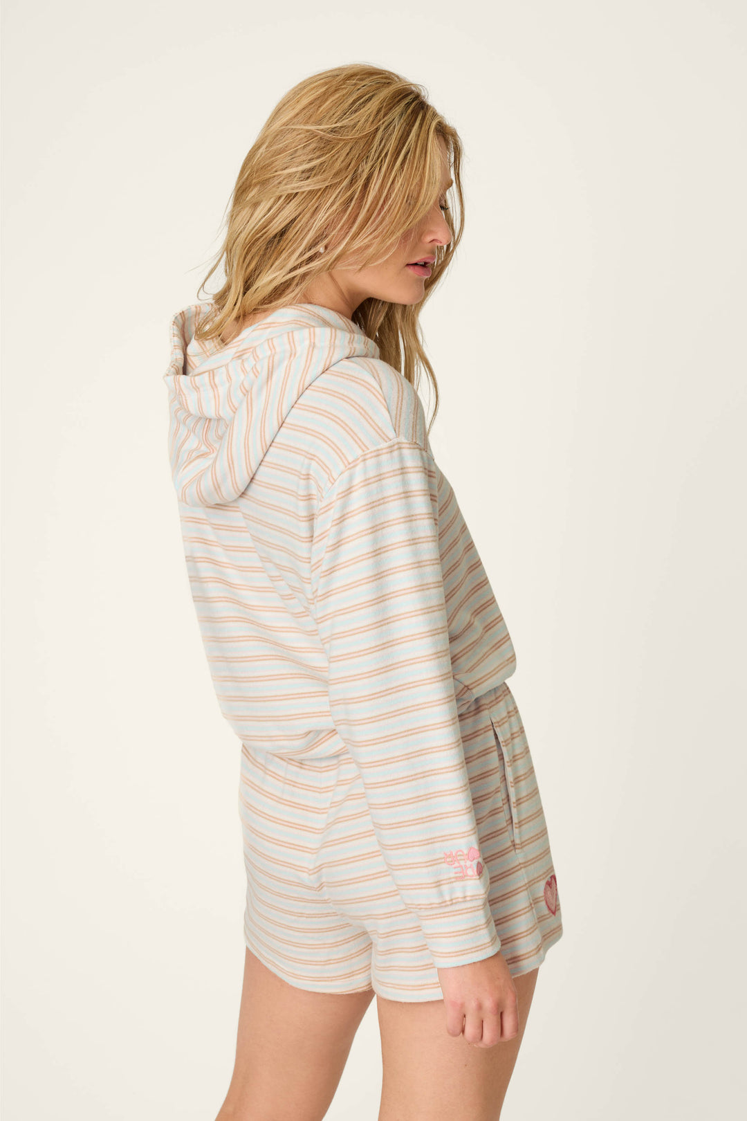 A person with long, wavy blonde hair wears the P.J. Salvage J'Adore Amour Sweetheart Stripes Hoodie and matching shorts, standing sideways and gazing slightly downward against a plain background.