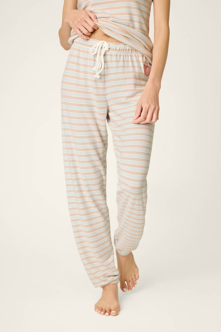A person wearing the J'Adore Amour Sweetheart Stripes Lounge Pants by P.J. Salvage, featuring pastel stripes, a drawstring waist, and a small heart patch on the hip, lifts their shirt slightly. They are barefoot against a plain background.