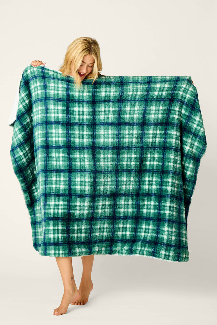 A person stands barefoot, smiling and admiring the P.J. Salvage Let’s Get Cozy Plaid Throw Blanket in green and blue, which they are holding up against a plain light-colored background.