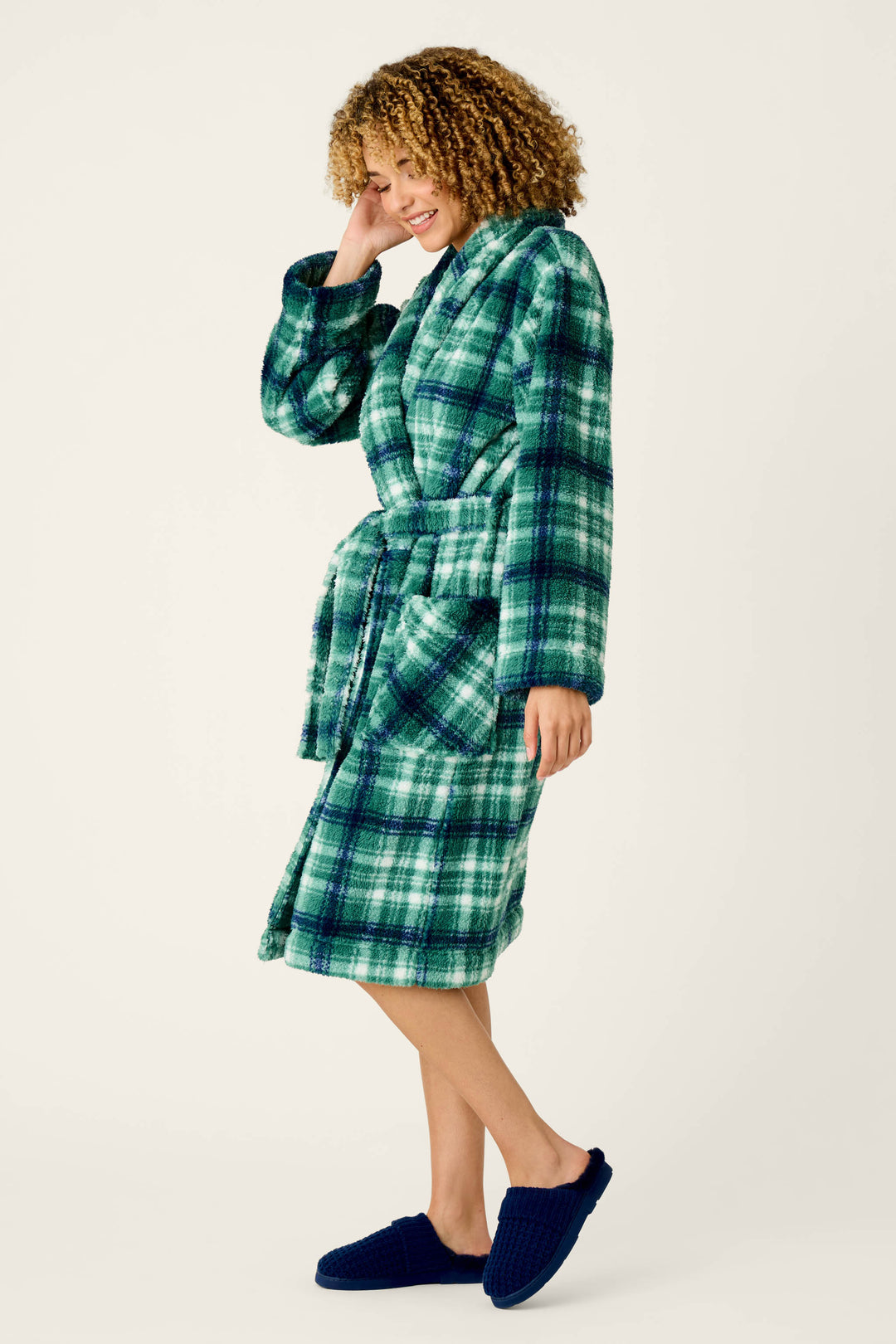 A person with curly hair stands smiling and posing against a plain light background, wearing the Let’s Get Cozy Plaid Robe by P.J. Salvage in a green and blue plaid pattern, paired with navy blue slippers.
