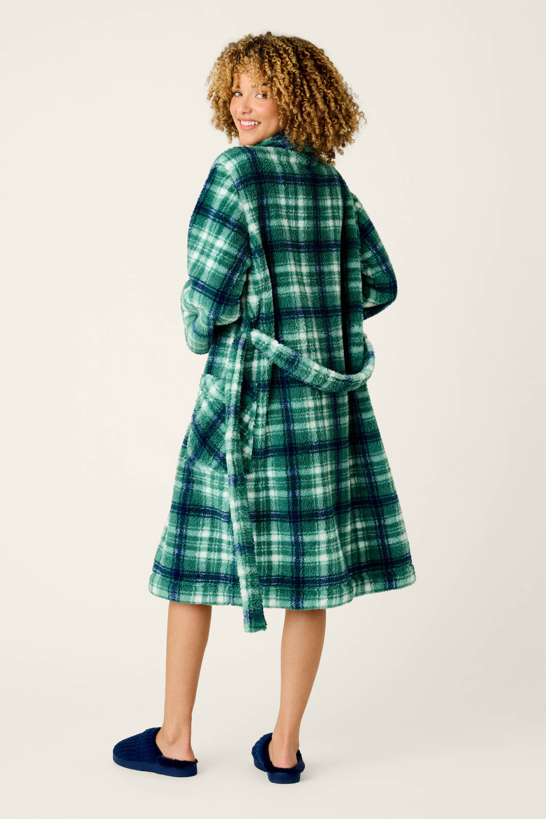 A person with curly hair wears the P.J. Salvage Let’s Get Cozy Plaid Robe in green and blue, paired with navy slippers, smiling as they look over their shoulder against a plain background.