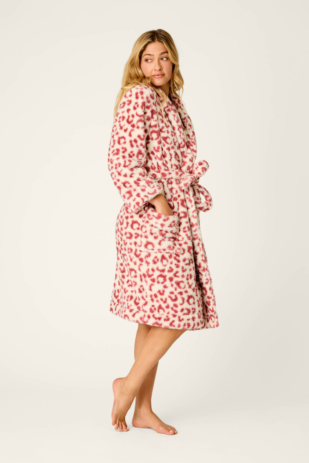 A woman stands barefoot against a plain light background, hands in the pockets of the P.J. Salvage Let’s Get Cozy Leopard Print Plush Robe, featuring pink and white animal print, as she looks to the side.