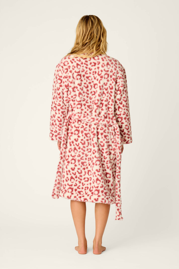 A person stands barefoot, facing away, wearing the P.J. Salvage Let’s Get Cozy Leopard Print Plush Robe—featuring a fuzzy pink and white leopard print and a belted waist. The background is plain and light-colored.