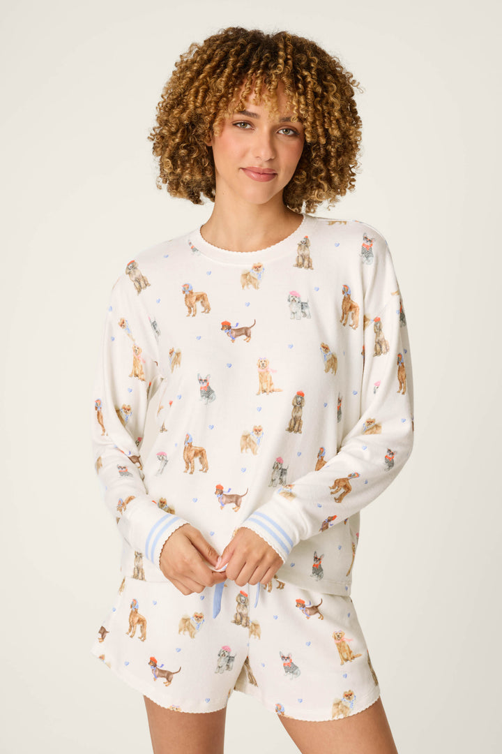 A woman with curly hair smiles gently while standing against a plain background, wearing the P.J. Salvage "Meet Me in Pawris" Dog Print Long Sleeve Top—a white pajama set adorned with colorful dog illustrations.