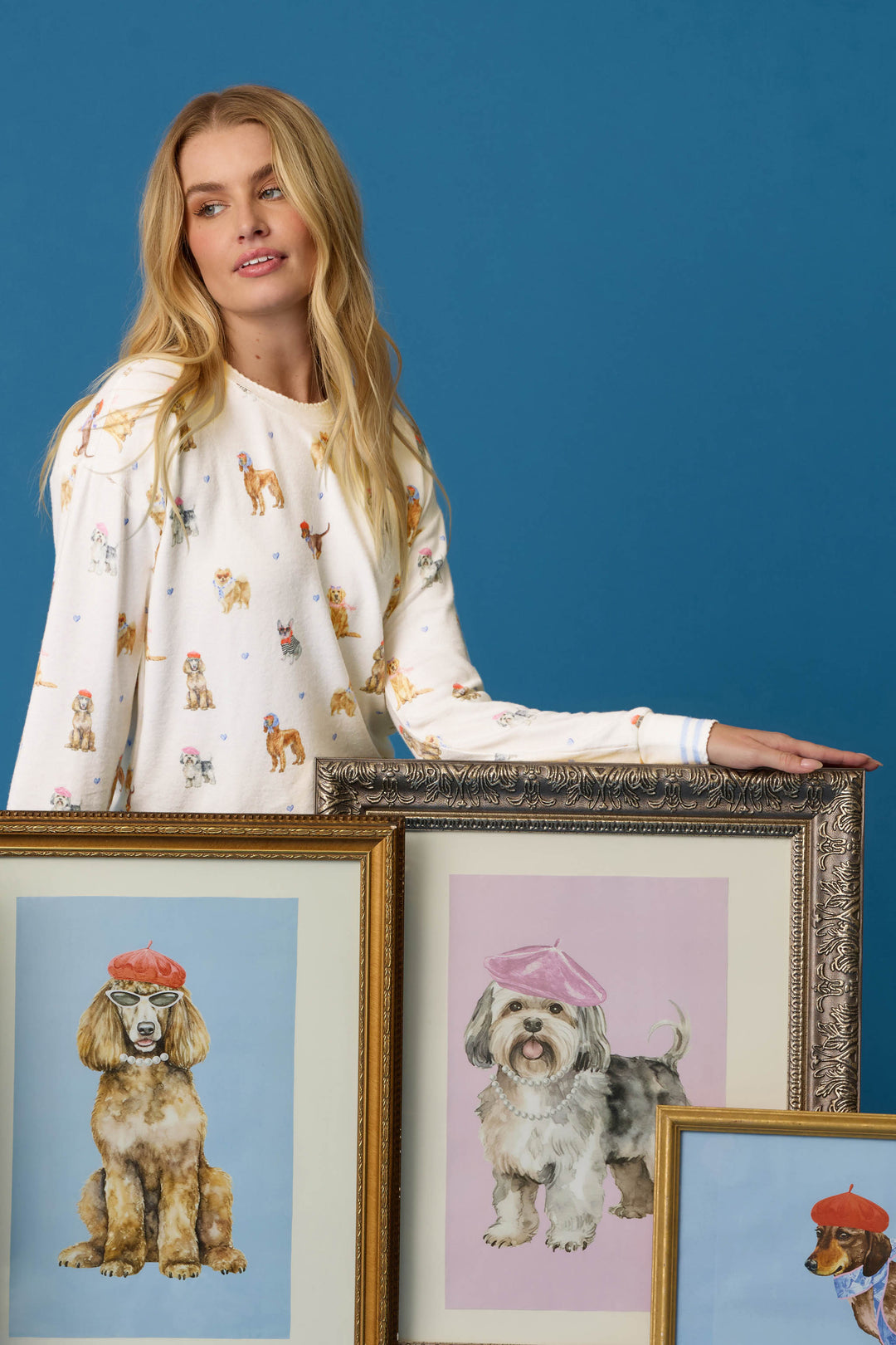 A woman wears the P.J. Salvage Meet Me in Pawris Dog Print Long Sleeve Top and stands beside three framed dog portraits—two featuring dogs in berets—against a blue background.