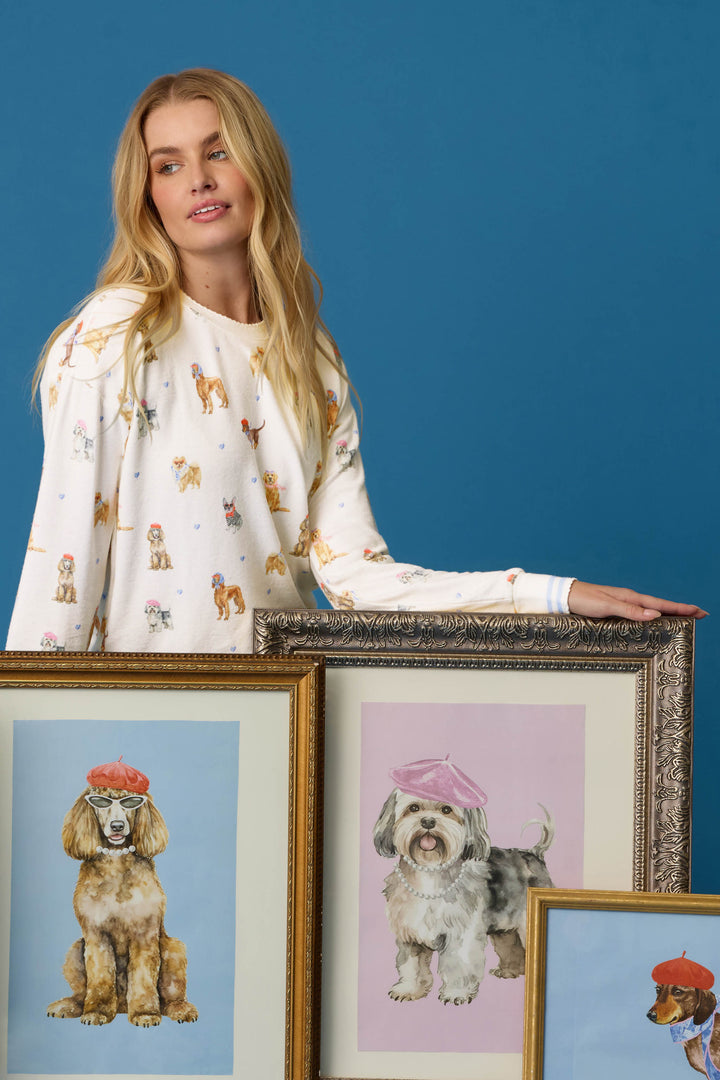 A woman wears the P.J. Salvage Meet Me in Pawris Dog Print Long Sleeve Top and stands beside three framed dog portraits—two featuring dogs in berets—against a blue background.