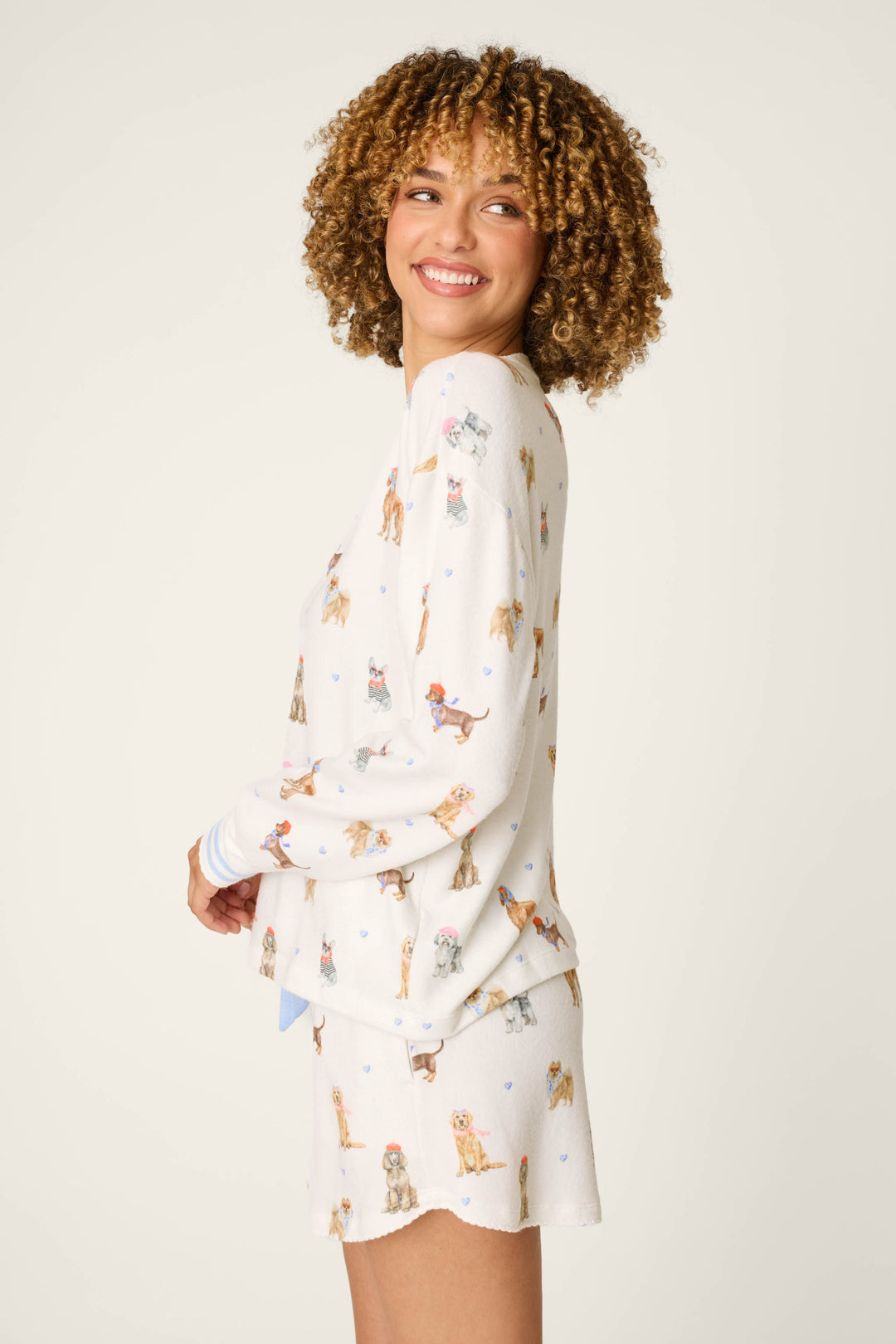 A woman with curly hair smiles as she stands against a neutral background, wearing the P.J. Salvage "Meet Me in Pawris" Dog Print Long Sleeve Top—part of a matching white pajama set adorned with colorful dog illustrations.
